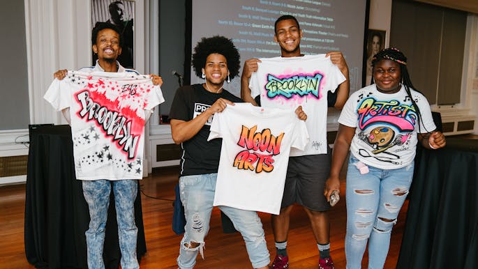 Four teens proudly display spray-painted white t-shirts with bright colors in the Weill Terrace Room.