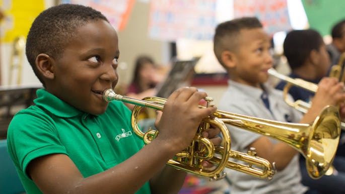 A small Black child with a bright green shirt smiles while playing the trumpet in a classroom.