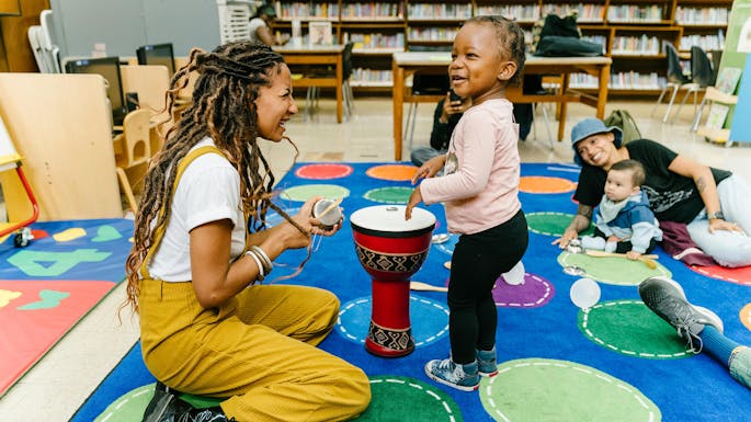 A young female teachers shows a toddler how to play a drum