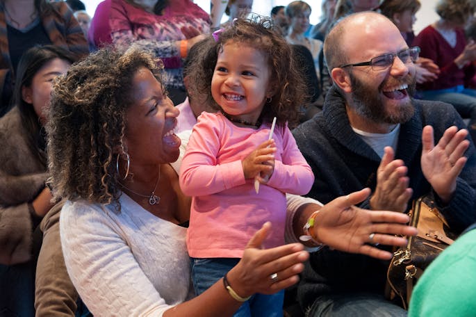 A Black woman smiles at a happy toddler close to her; a White man with glasses laughs nearby.