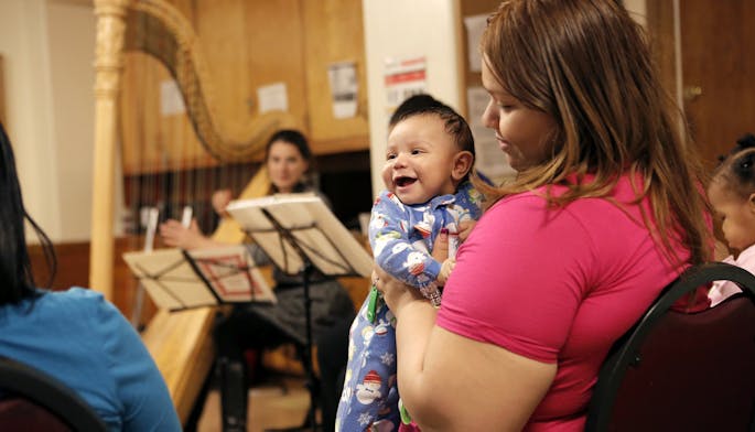 A woman in a pink shirt looks adoringly at a toddler in a onesie; a harpist sits in the background.