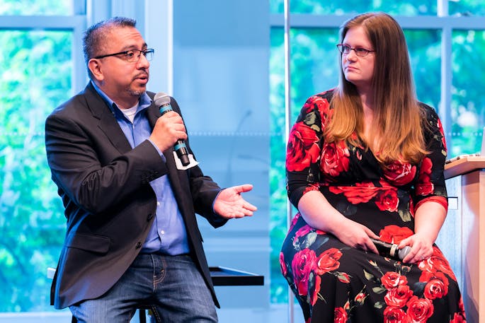 A seated man with glasses speaks into a microphone; a woman in a floral dress listens to him.