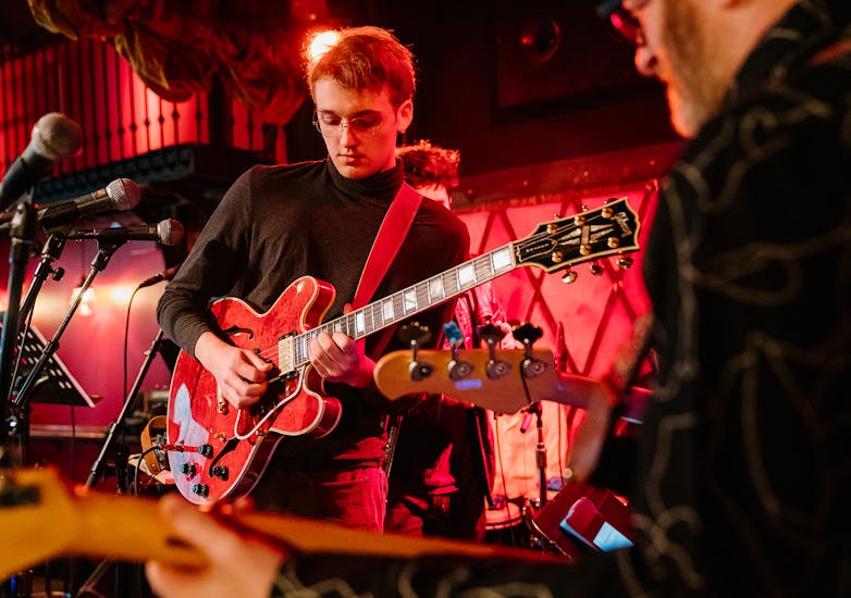 A teenage boy in a turtleneck and glasses plays a red guitar with a red spotlight filling the club.