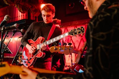 A teenage boy in a turtleneck and glasses plays a red guitar with a red spotlight filling the club.