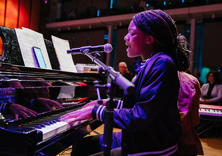 A teenage girl with gathered braids in a dark jacket sings into a microphone and plays the piano.