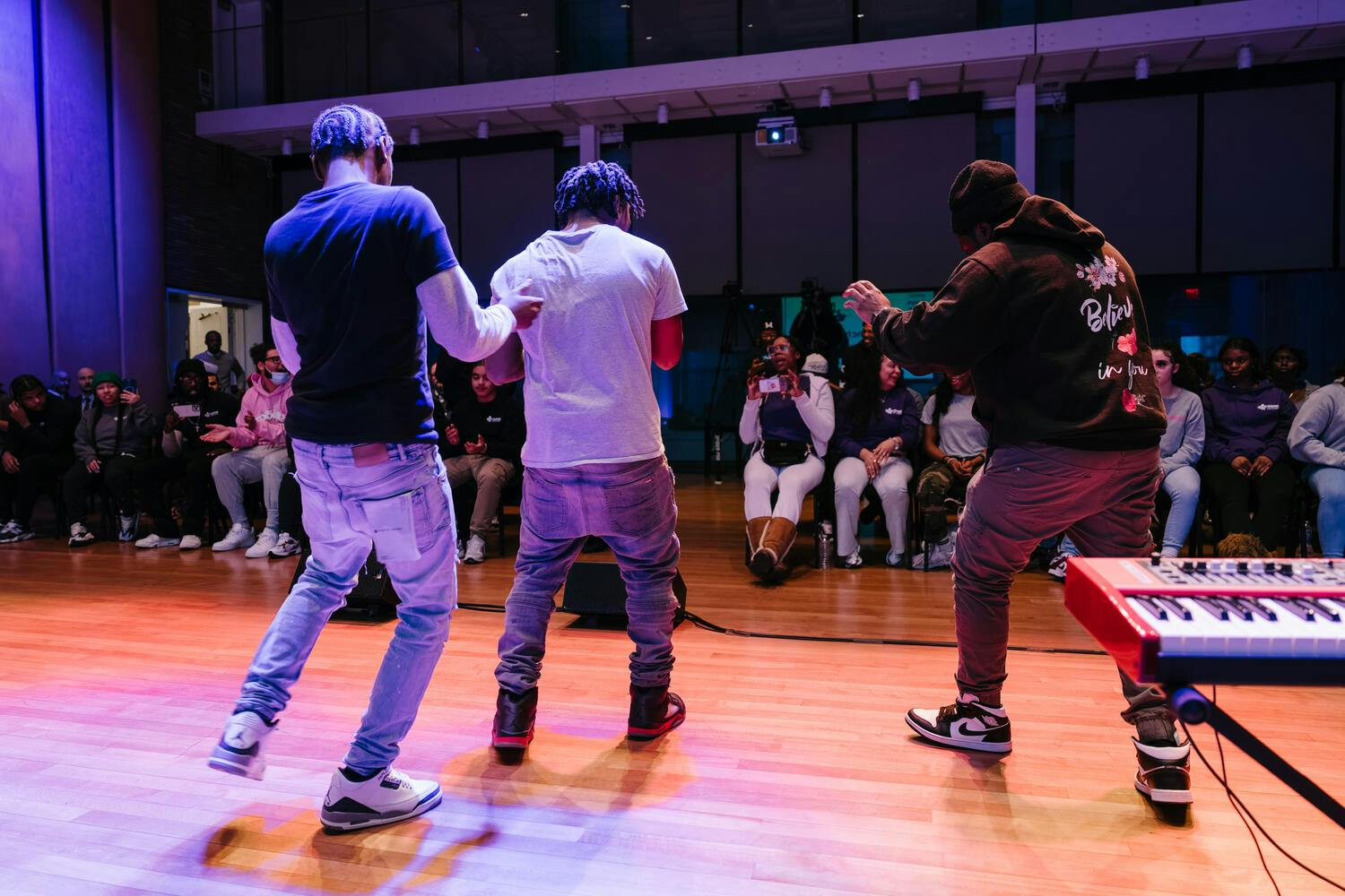 Three young men wearing jeans and t-shirts dance before a small audience