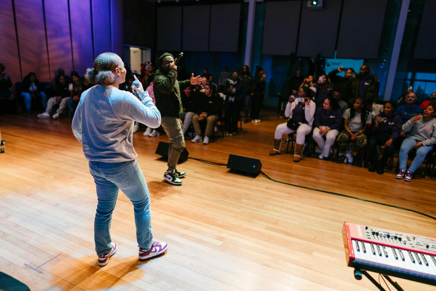A young woman and man wearing jeans and sweatshirts sing before a small audience