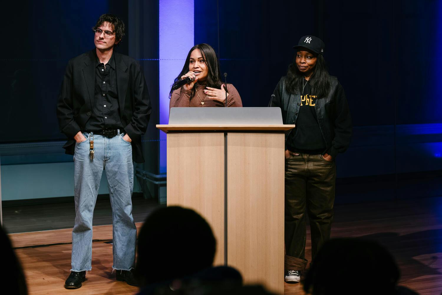 A young woman speaks from a podium, flanked by a young man and woman