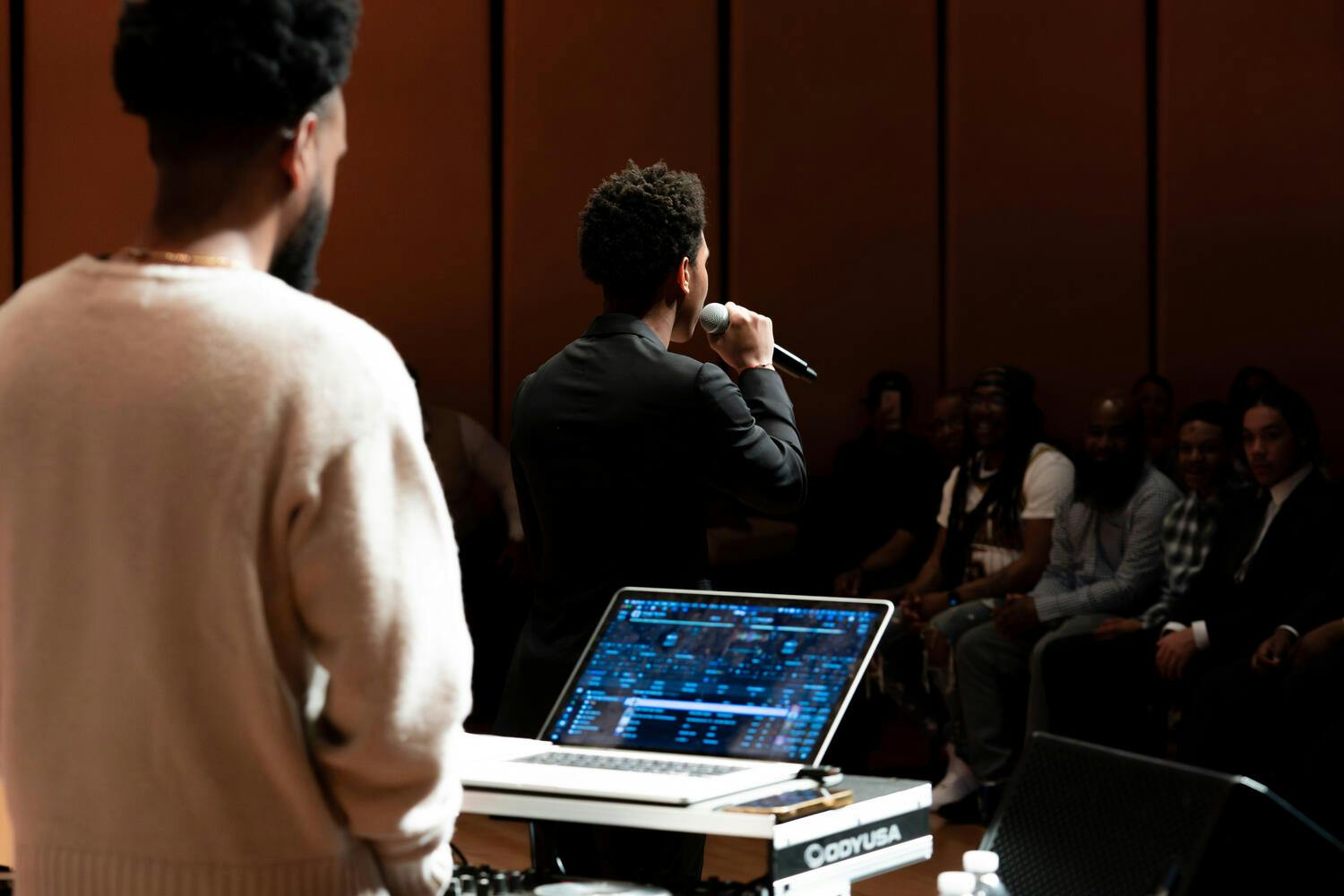 One man stands at mixing program on his PC as another speaks into a mic before an audience