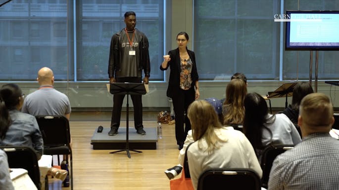 A man at a music stand listens as a presenter guides a class of teachers