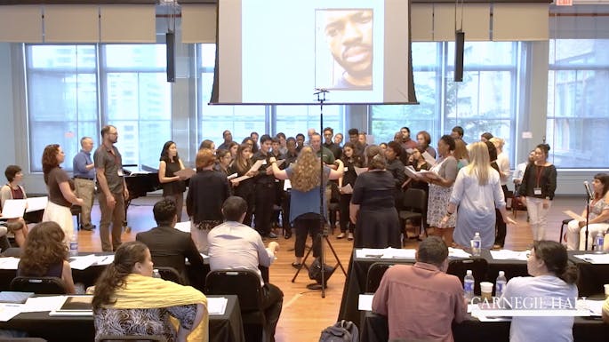 A presenter conducts a choir of teachers as an audience of teachers watches