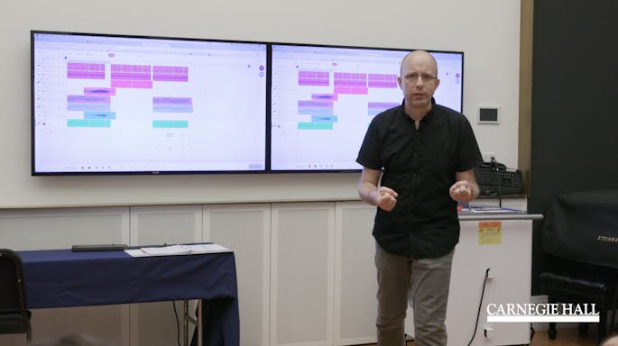 A presenter speaks as two monitors behind him display graphs