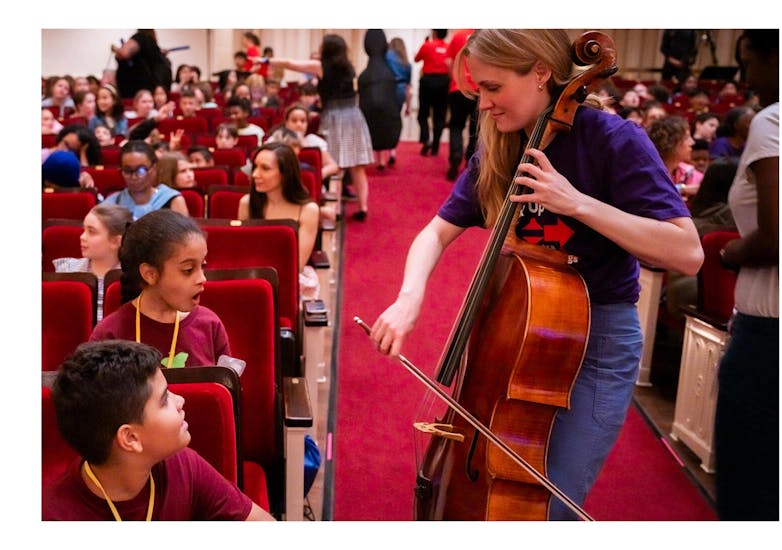A boy and girl seated in the audience at Carnegie Hall watch goggle-eyed as a woman plays cello for them