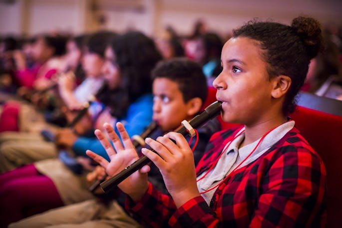 A young girl in a red flannel shirt plays the recorder with other kids playing in the background.