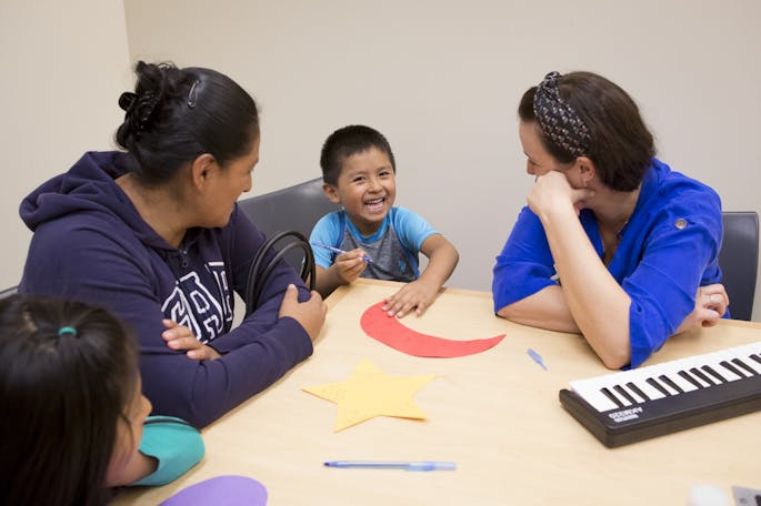 Two women in blue tops look at a smiling small boy behind a paper crescent moon.