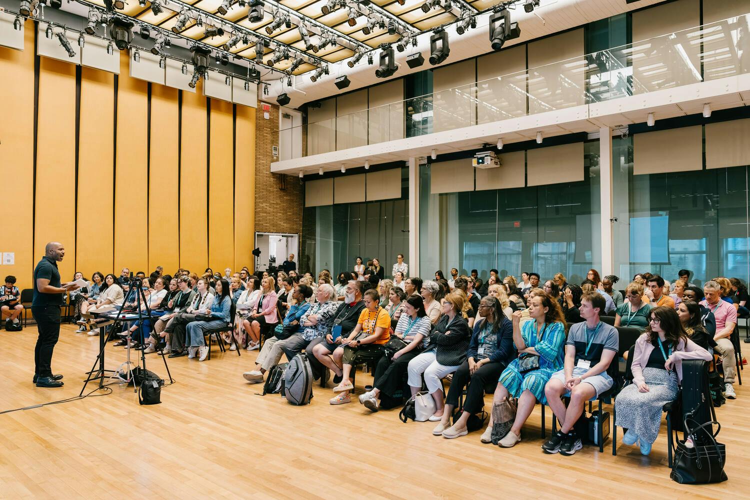 A man speaks before an audience in a wood-paneled rehearsal studio
