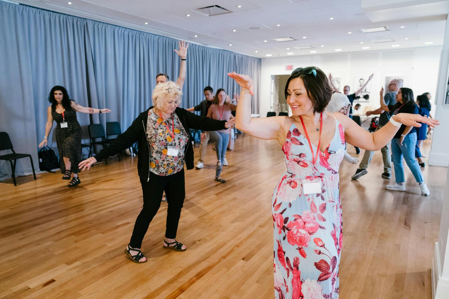 A young woman wearing a floral dress leads a group of young and elderly women in a dance