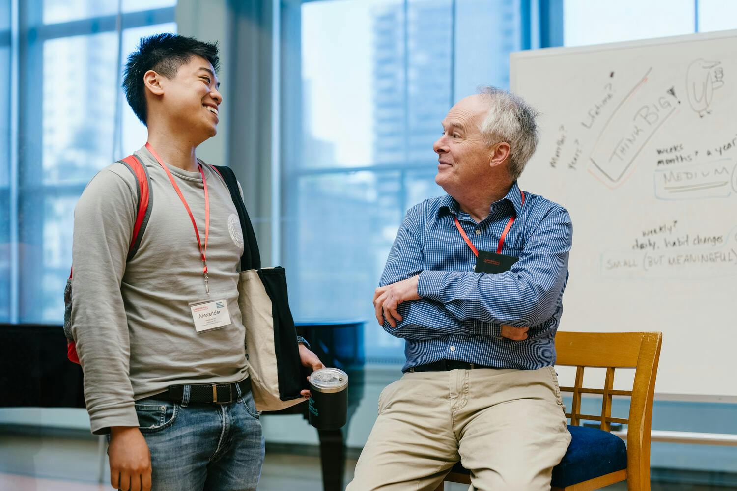 An older man talks with a smiling young man, a whiteboard behind them