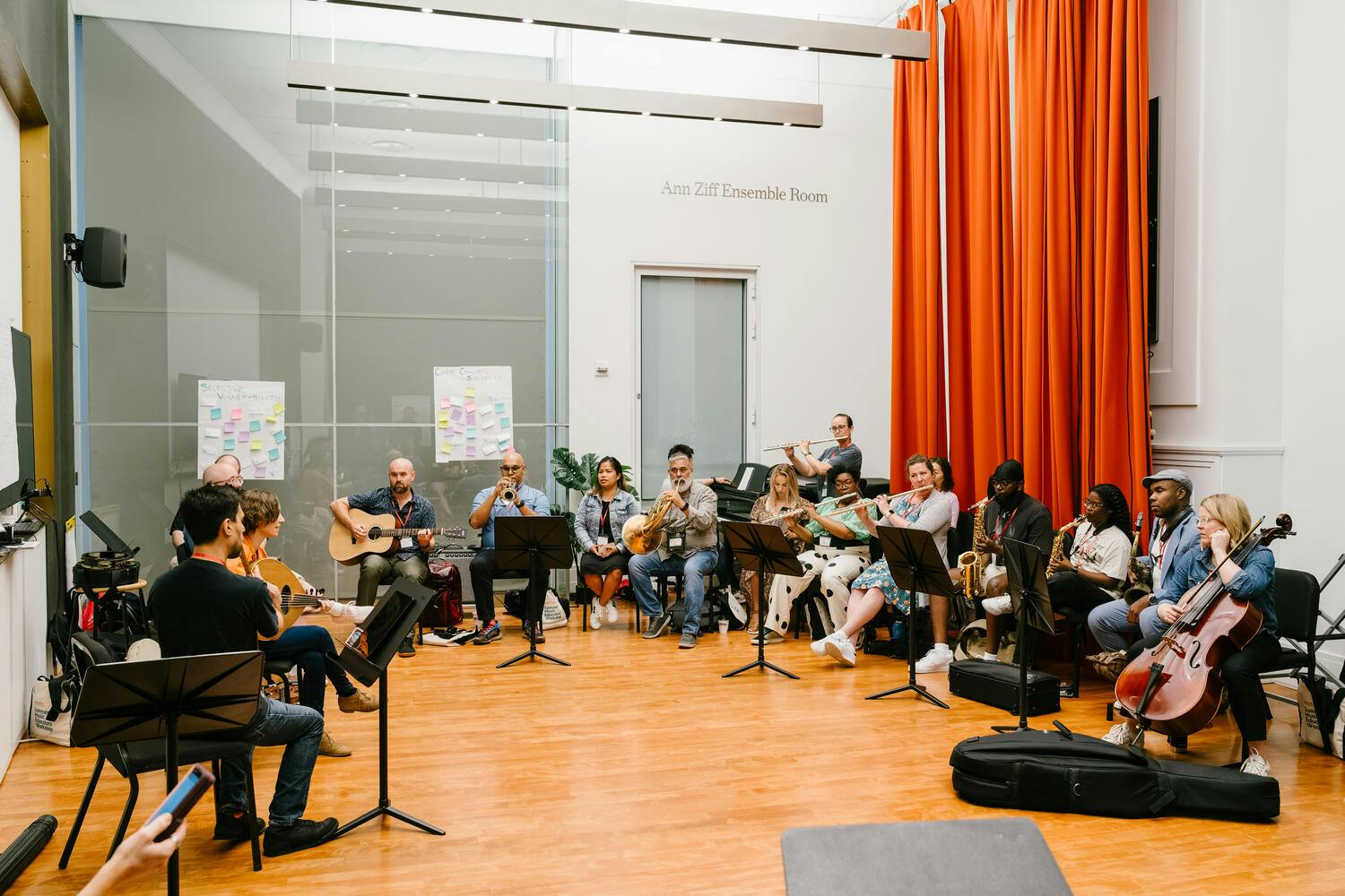A group of musicians play their instruments, sitting in a semi-circle in a rehearsal room