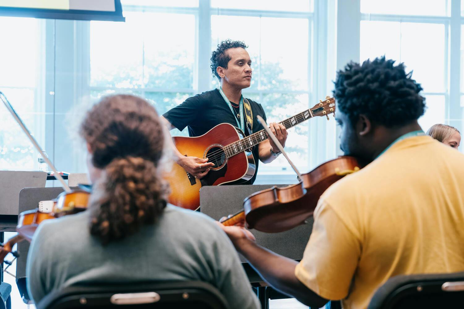 A man plays guitar before a man and woman seated who are playing violins