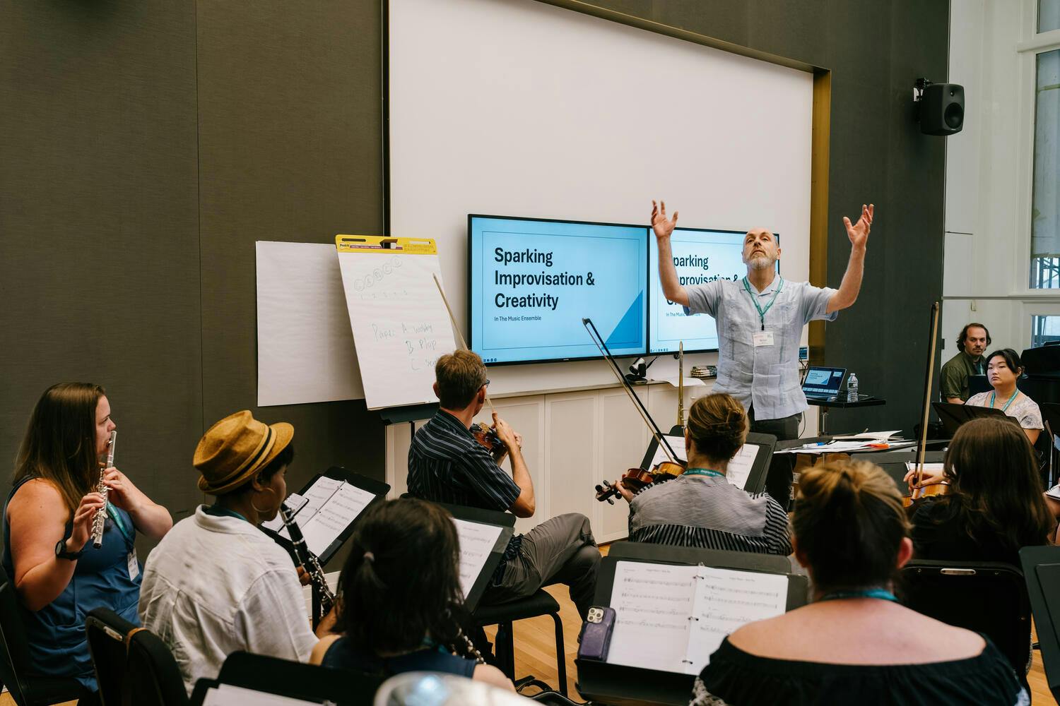 A man gives a presentation on "Sparking Improvisation and Creativity" as men and women watch, holding their musical instruments