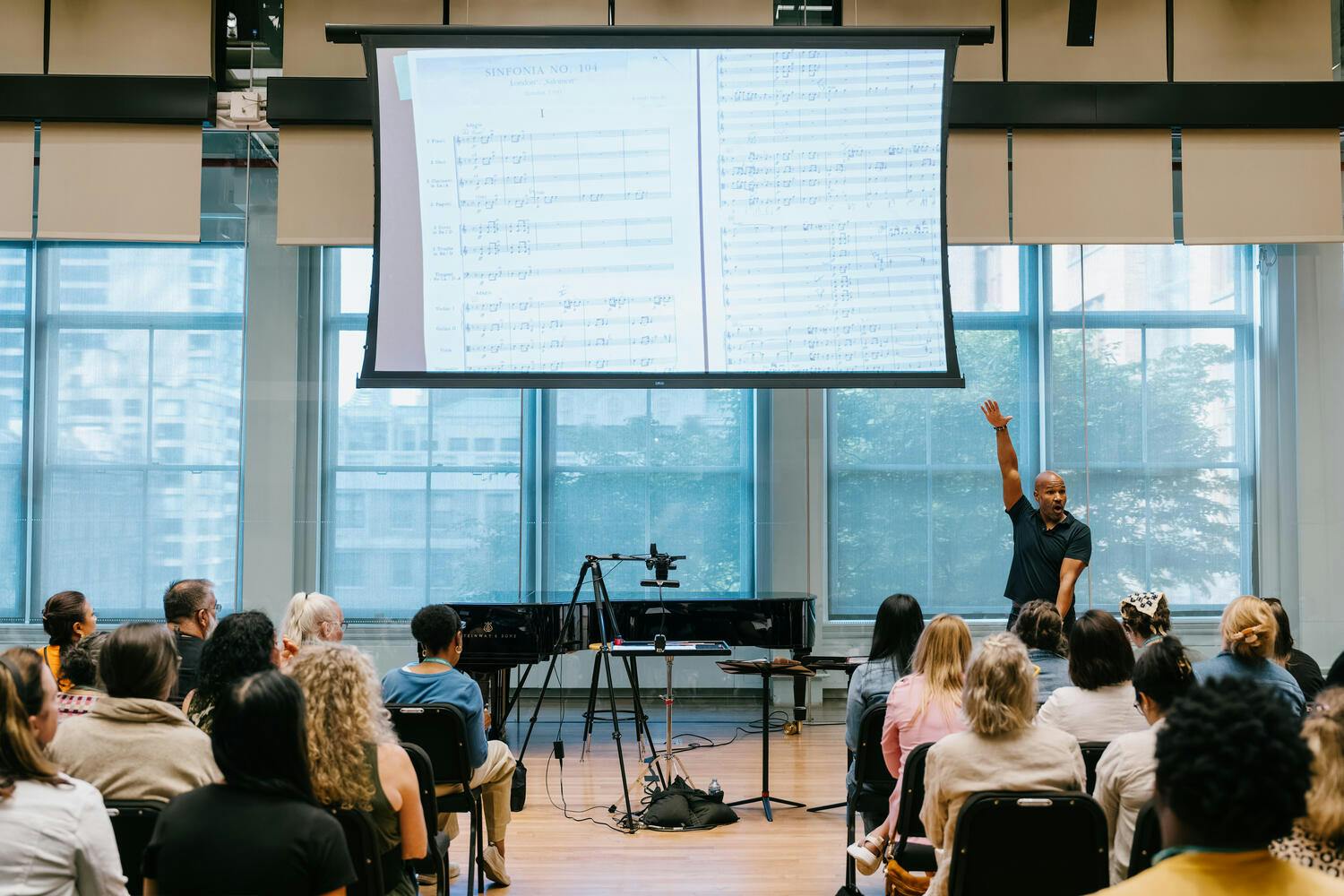A man stands with arms uplifted before an audience, sheet music projected on a screen