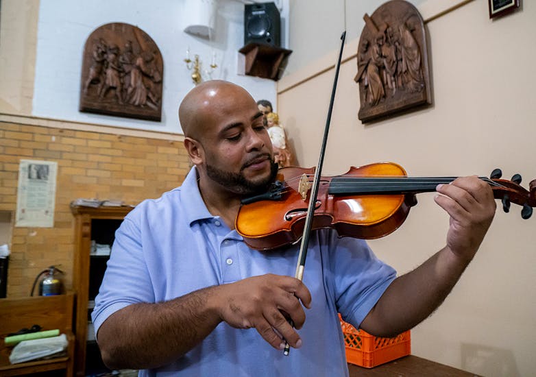 A bald man with a beard wearing a blue polo plays the violin in a chapel.