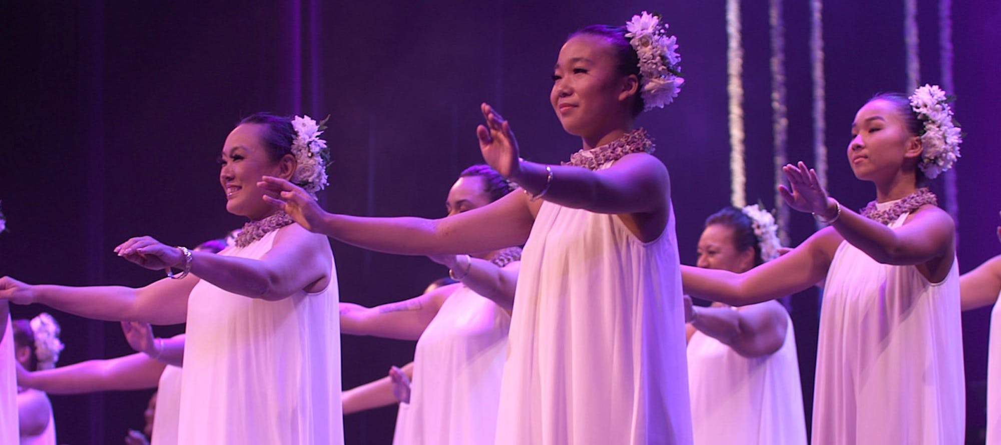 Young women in flowing white gowns with flowers in their hair hold their arms out