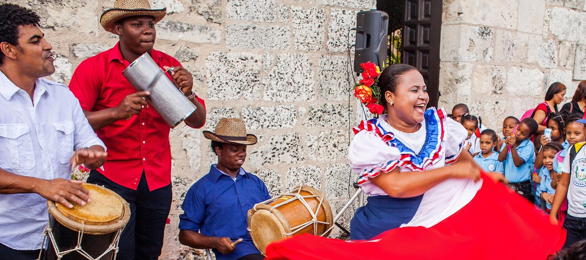A woman in a blue and red dress dances as two men play percussion instruments and a crowd of children watch