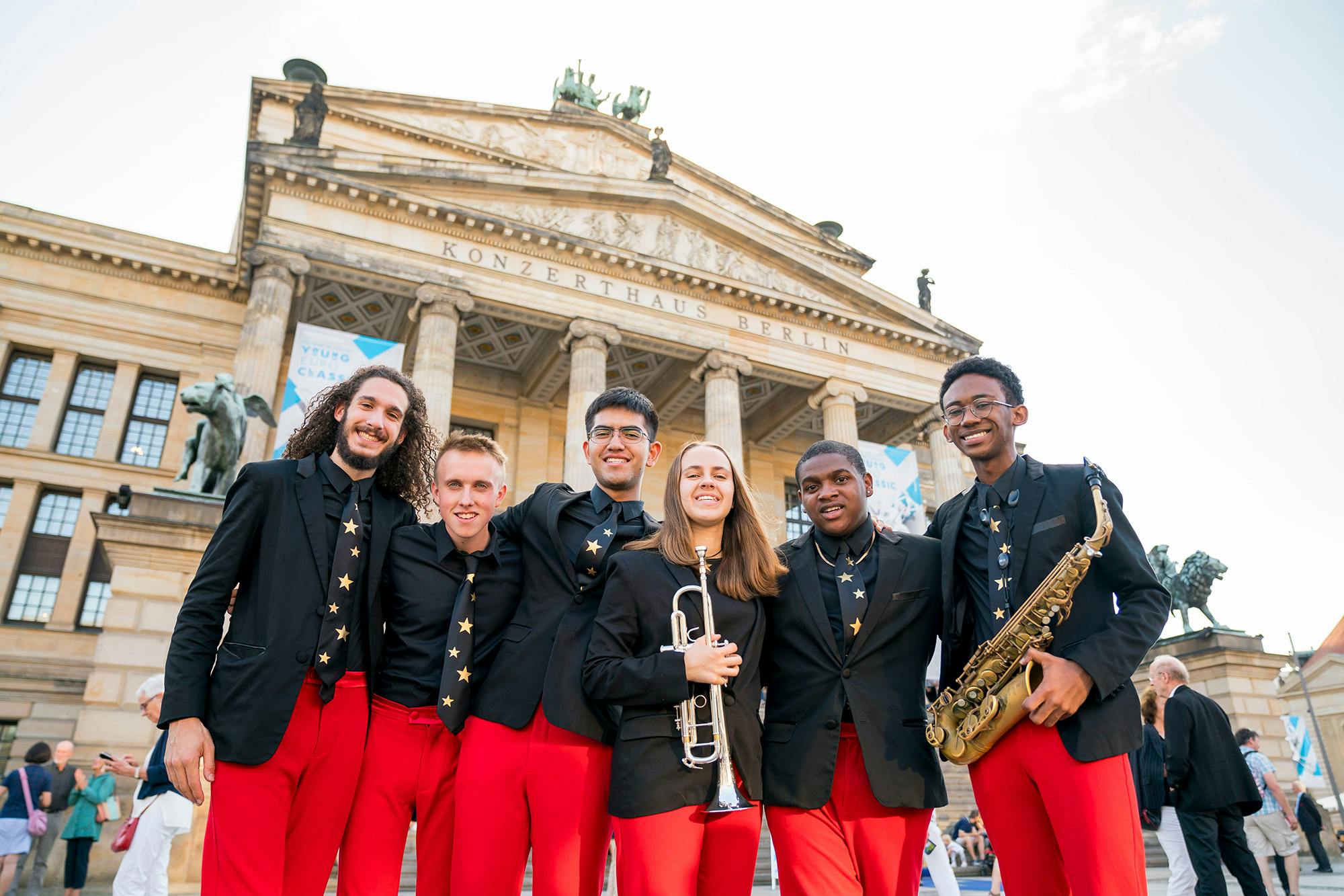 NYO Jazz musicians wearing black blazers and red pants stand together outside the Konzerthaus Berlin.