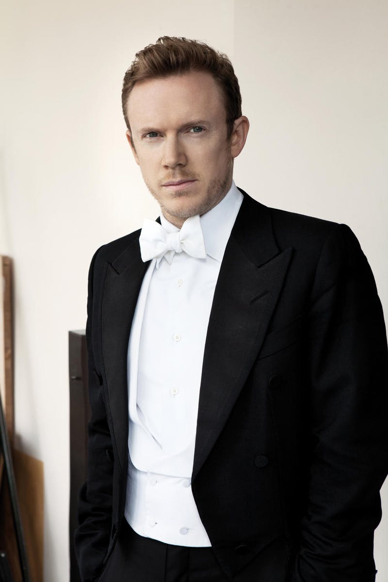 A blond-haired man with a moustache wearing a tuxedo looks into the camera