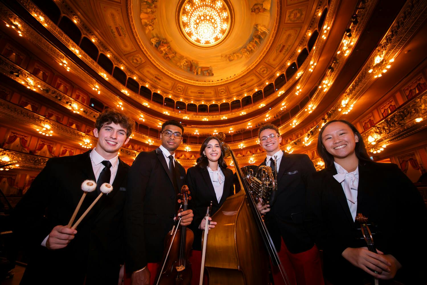 Five male and female NYO-USA musicians pose together in an ornate auditorium, chandelier overhead