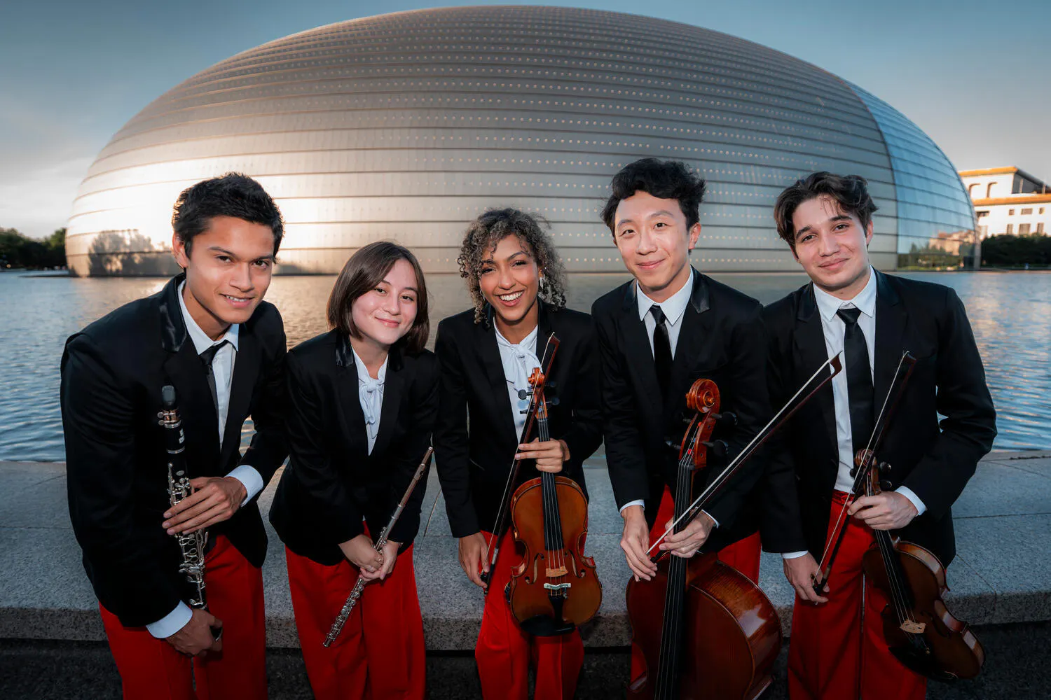 Five NYO-USA musicians wearing red pants and black blazers pose with their instruments before a gold dome-shaped performance hall