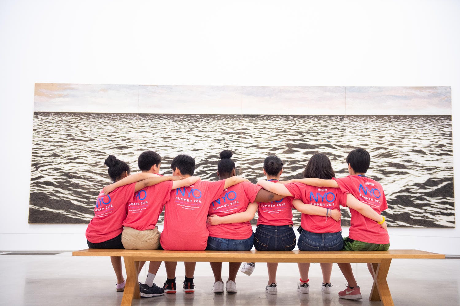 Musicians of NYO2 sit arm-in-arm before an image of the ocean in an art museum 