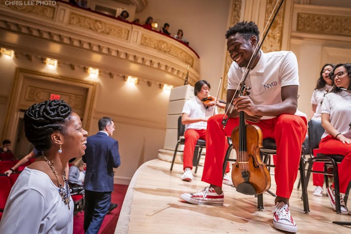 An NYO2 violinist wearing a white polo and red pants smiles at an audience member in Carnegie Hall.