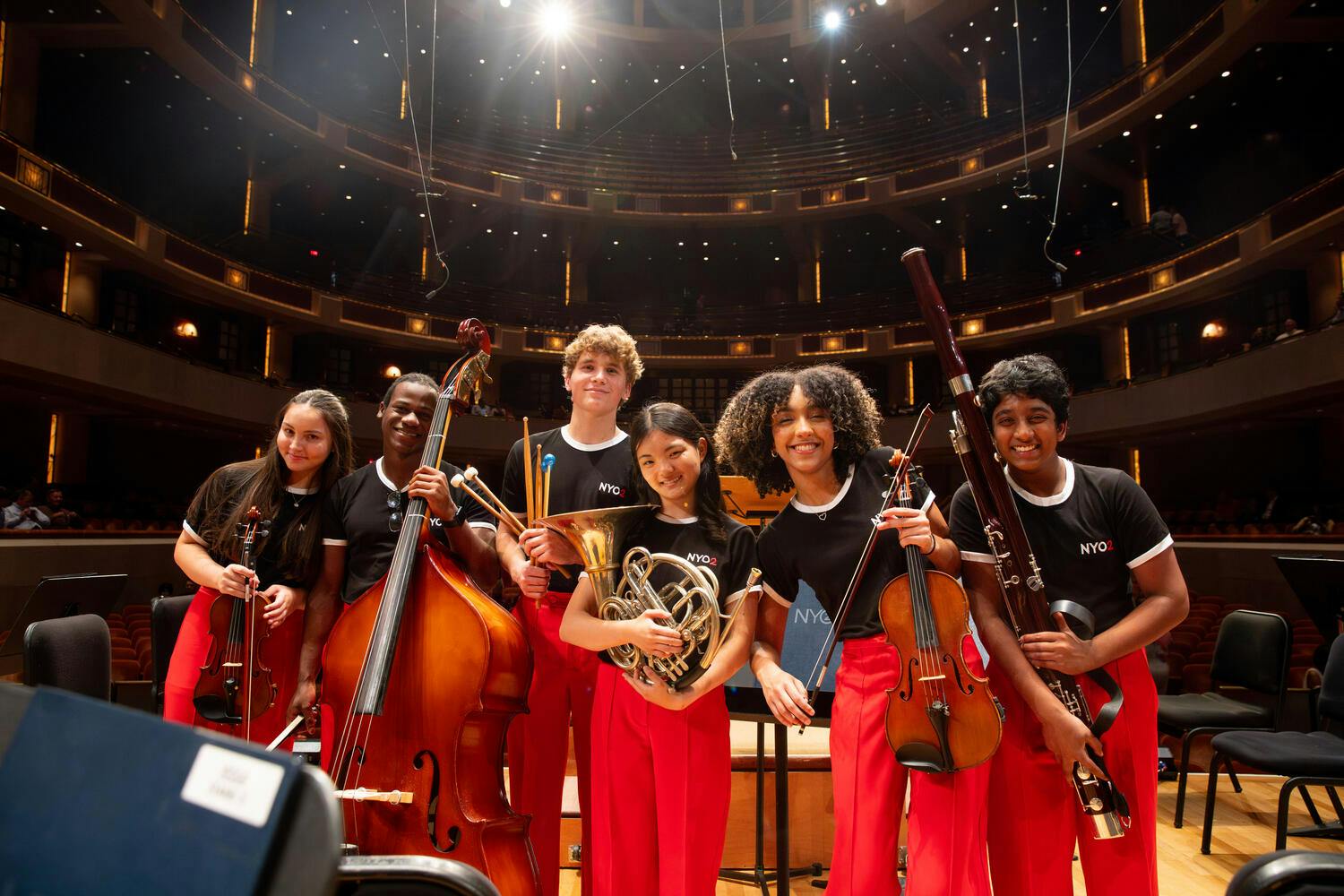 NYO2 musicians wearing red pants and black t-shirts pose with their instruments on stage at Carnegie Hall