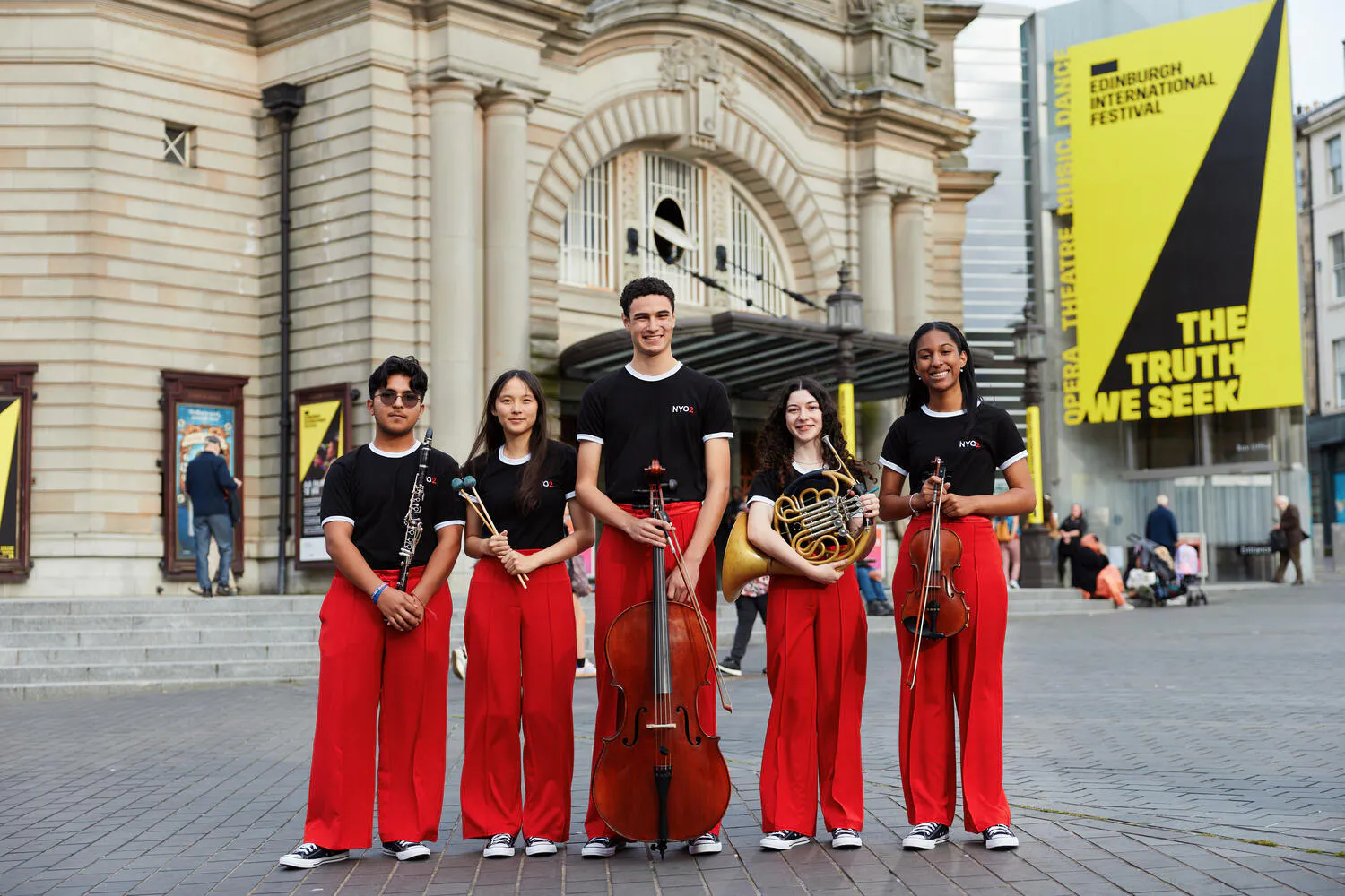 Five NYO2 musicians wearing red pants and black t-shirts pose with their instruments outside a performance hall