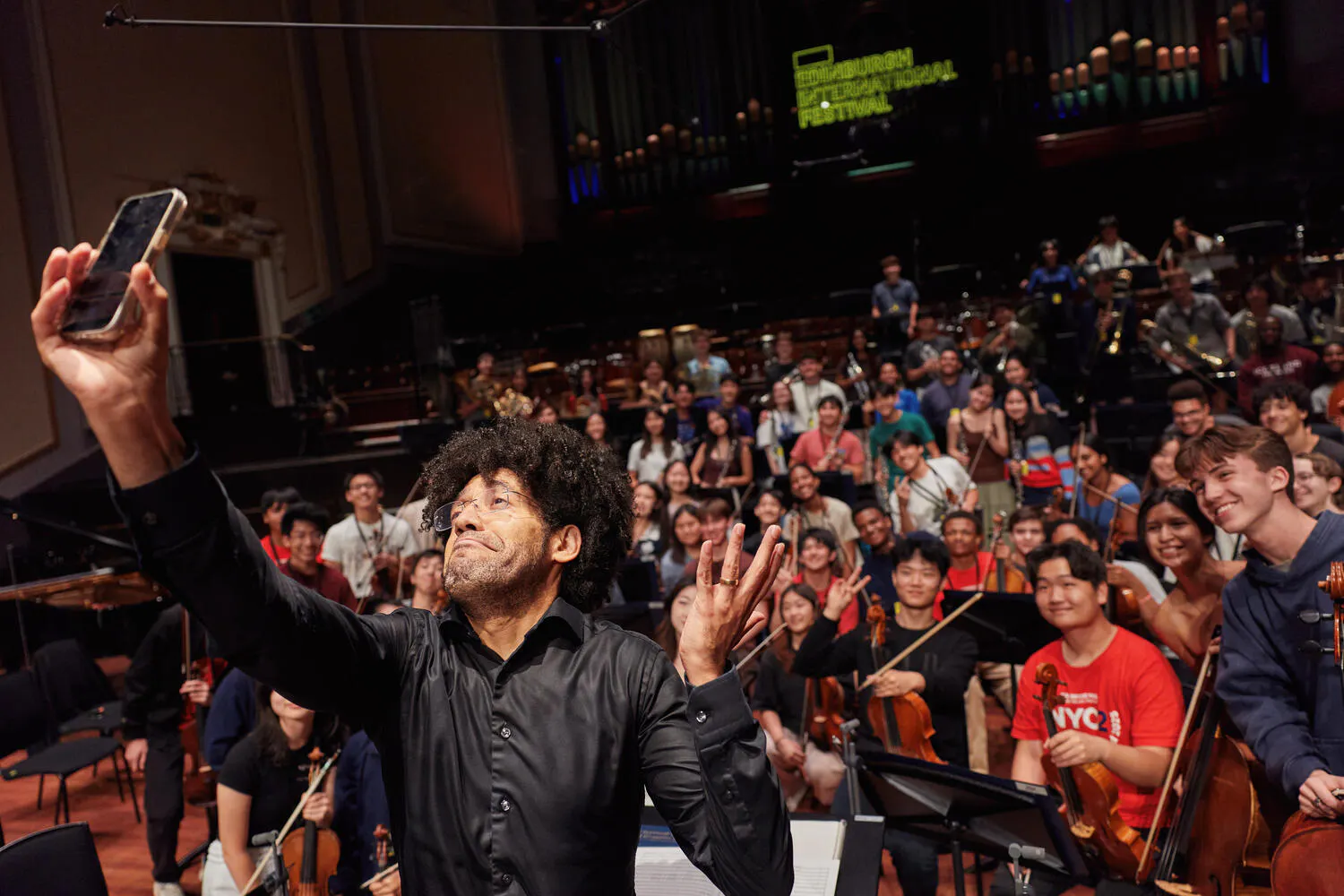 A man takes a selfie of himself with the NYO2 Orchestra behind him on stage