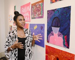 A woman stands in front of a wall of colorful paintings.