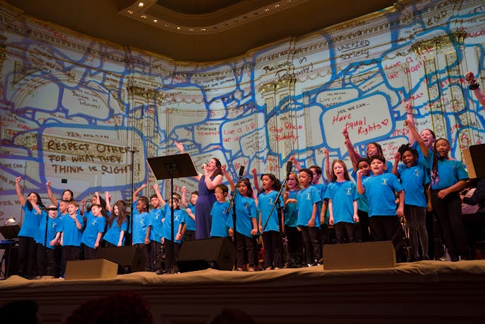 A choir of children sing with inspirational hopes projected behind them.