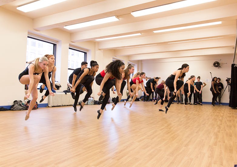 A group of dancers—snapping, as they jump mid-air—rehearse in a studio.