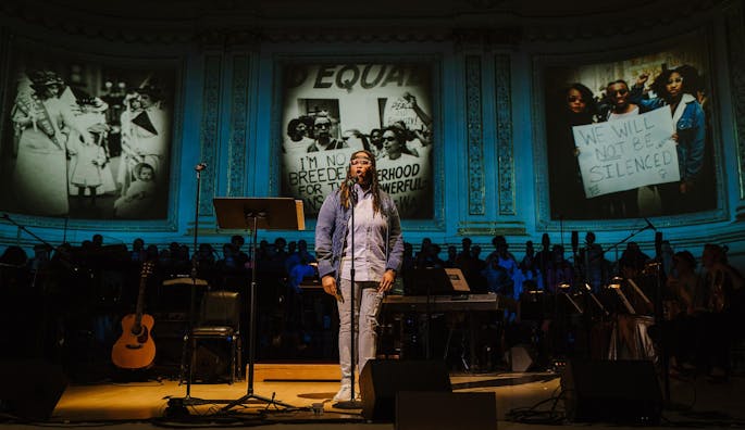 A Black, female poet onstage surrounded by musicians and photos of the Civil Rights movement
