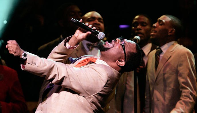 A Black, male singer is bent over backwards while singing with his bandmates.