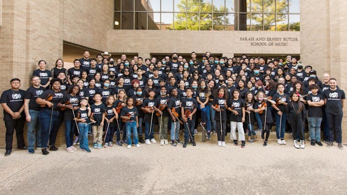 A group of teachers and students holding instruments in matching t-shirts poses outside a school.