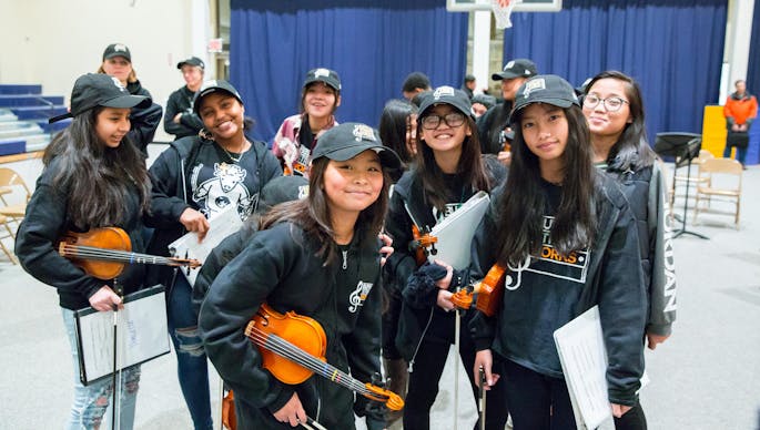 A group of girls wearing black caps and dark clothing hold violins and music in an auditorium.