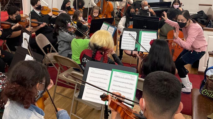 A woman in a pink sweater leads a rehearsal of children playing instruments.