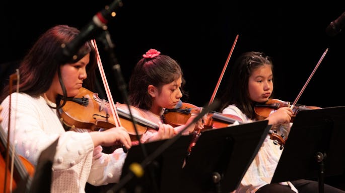 Three girls play the violin seated, wearing white blouses.