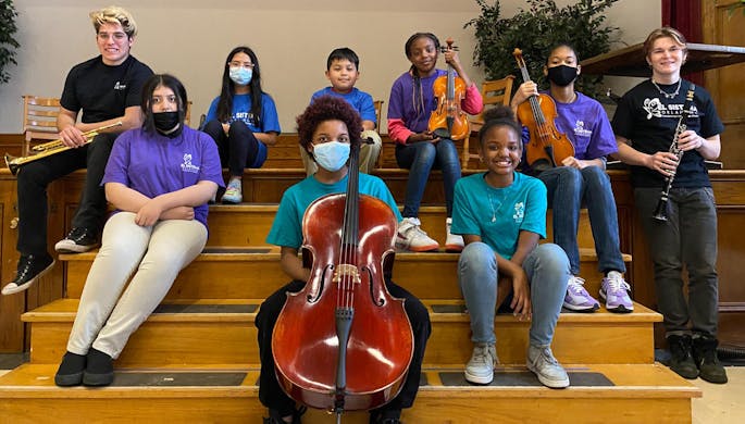 Kids and teens sit on steps, smiling as they hold a variety of instruments.