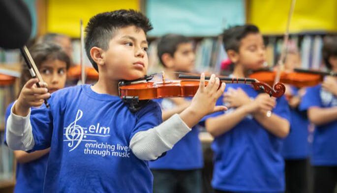 A young boy with short black hair wearing a blue ELM t-shirt holds a violin under his chin.