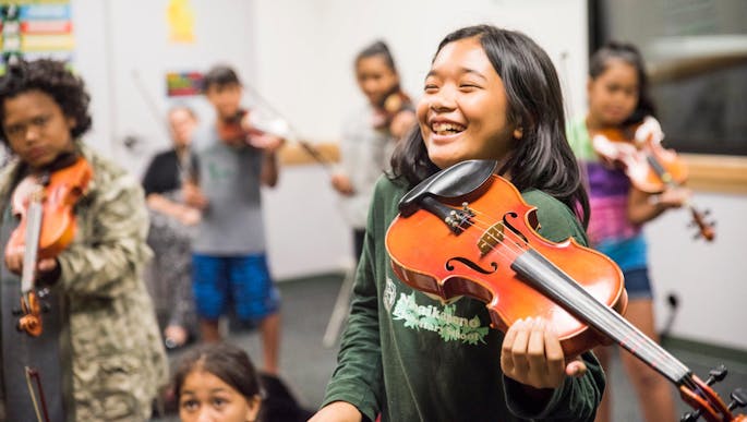 A smiling kid with brown hair wearing a long sleeve green t-shirt, smiles and holds a violin.